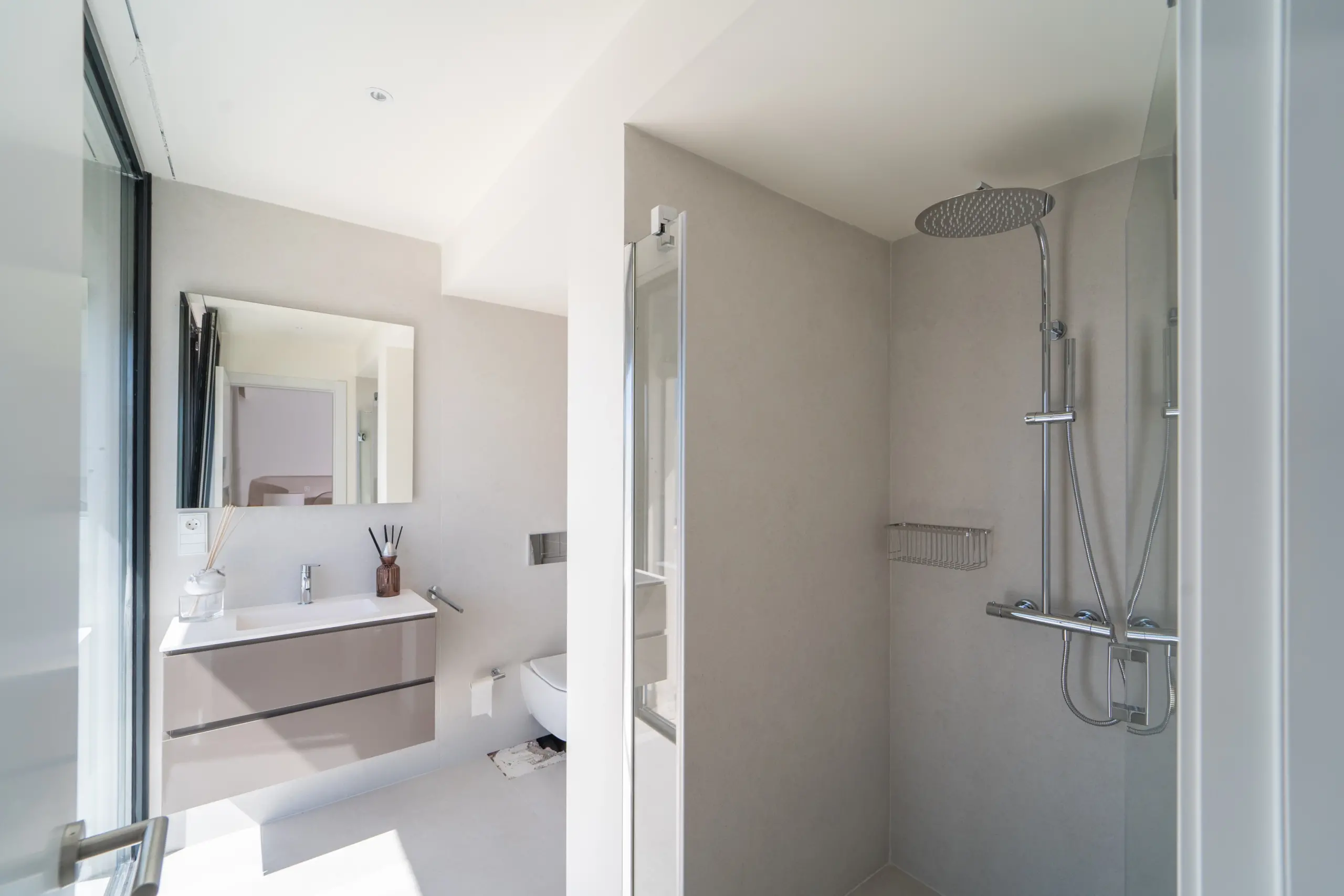 Interior de un baño moderno y luminoso, equipado con una ducha de cabezal grande, espejo grande que refleja una vista parcial del dormitorio contiguo y un lavamanos con accesorios modernos. Diseño simple y colores neutros dominan el espacio.