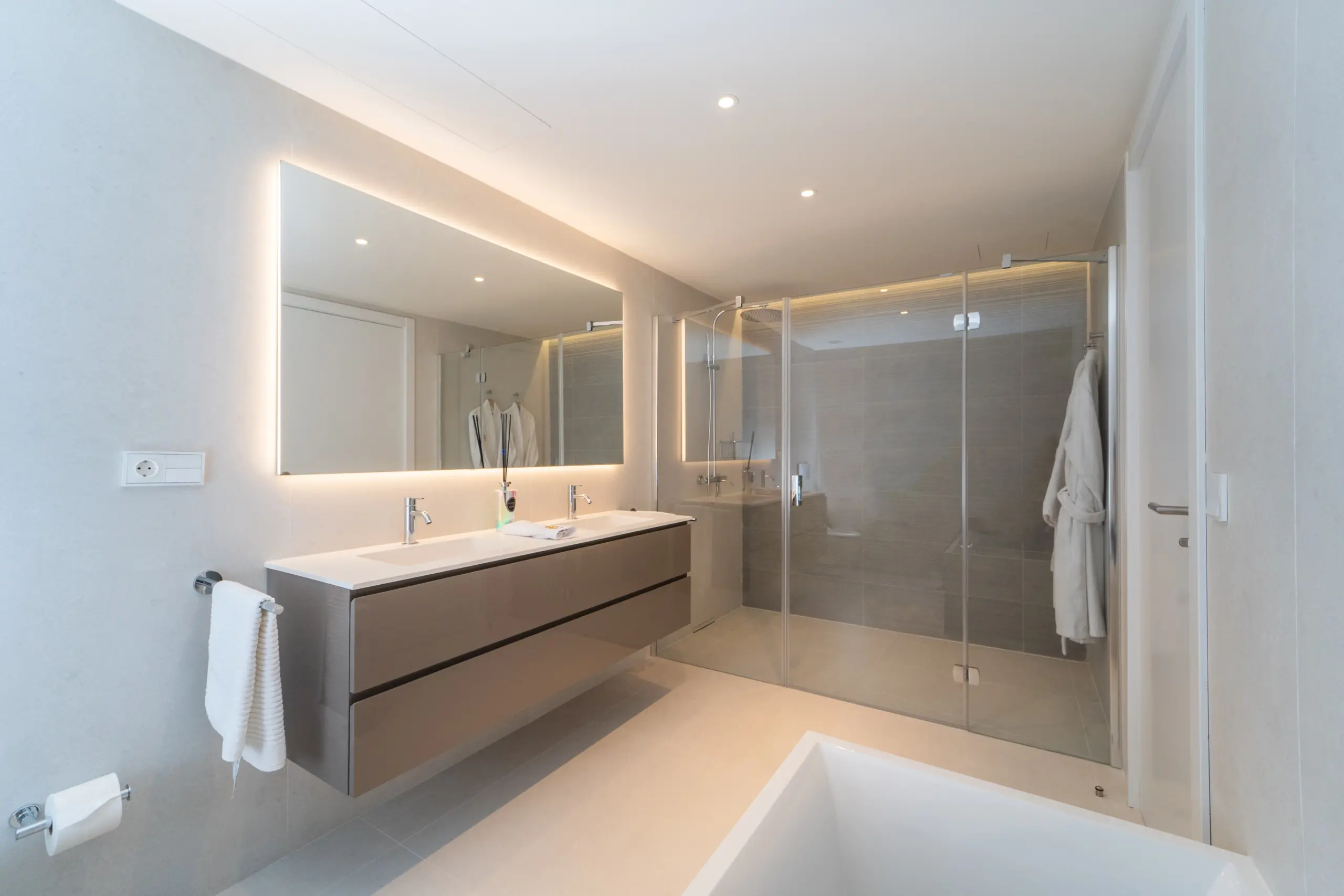 Baño principal de lujo con dos lavamanos, espejos grandes y una ducha amplia de vidrio. Iluminación LED alrededor del espejo principal añade un toque sofisticado. Decorado en tonos beige y grises, transmitiendo un ambiente tranquilo y elegante.