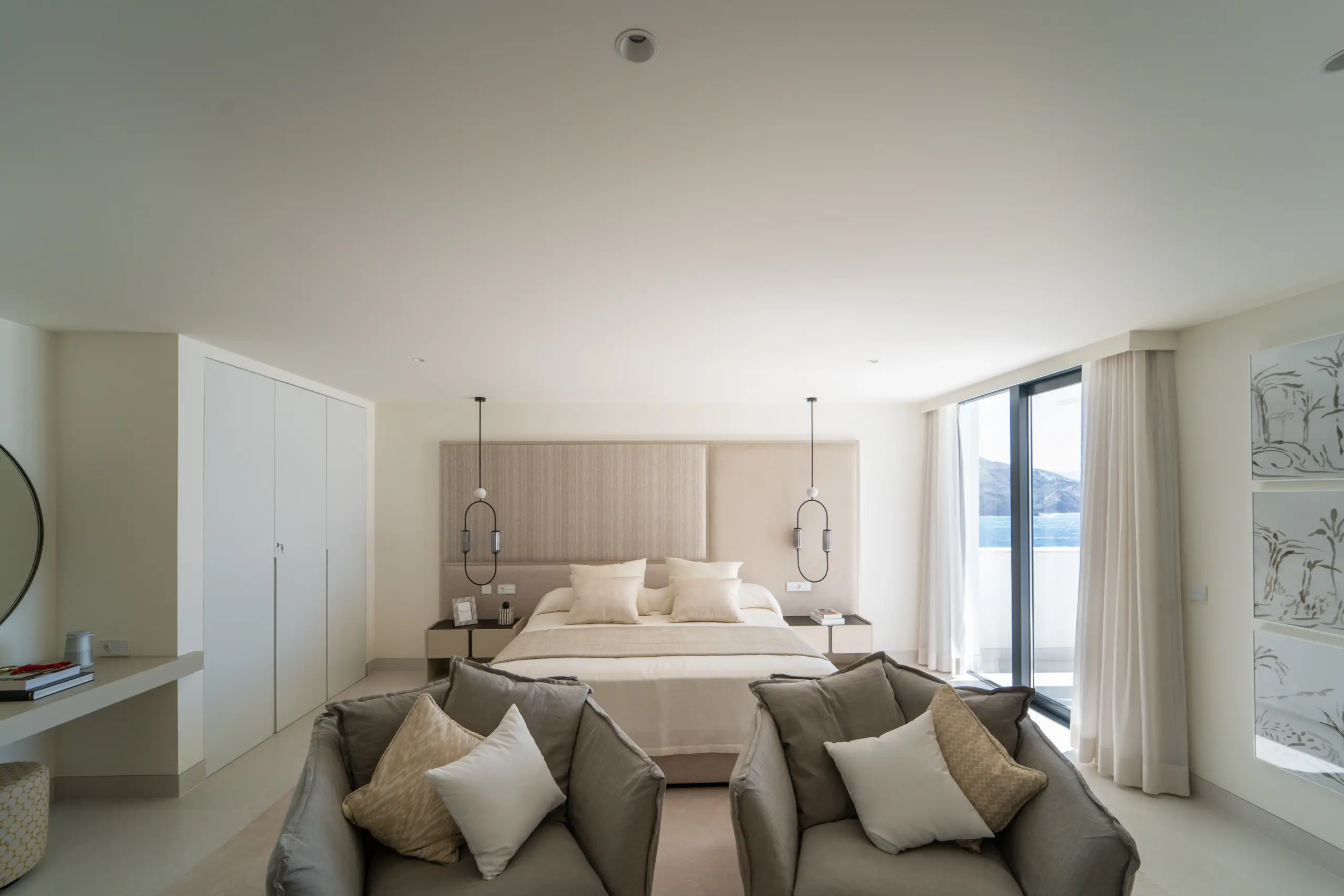 Dormitorio espacioso y moderno con una cama grande en el centro, rodeada de dos sillas cómodas en un área de descanso con cojines. Decoración minimalista en tonos beige y blancos. Grandes ventanales ofrecen una vista impresionante del mar y montañas al fondo.